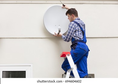 Young Male Technician In Uniform Installing TV Satellite Dish On Wall