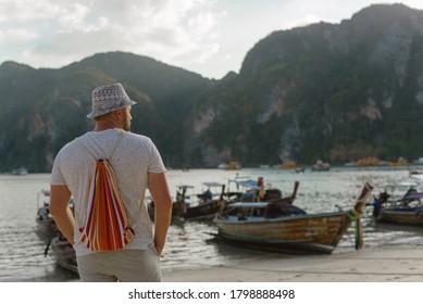 Young male tourist in a hat and with a backpack on a Thai beach on a background of traditional Thai wooden boats.