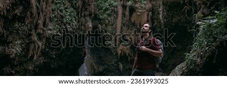 A young male tourist stands near a tropical waterfall among the rocks.  A man, a traveler with a backpack in the tropical jungle in the mountains overgrown with plants