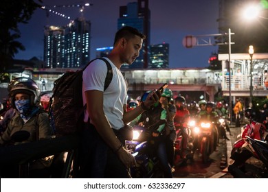 Young Male Tourist Using The Mobile Phone While Walking On A Crowded Street With People On Scooters In Jakarta At Night