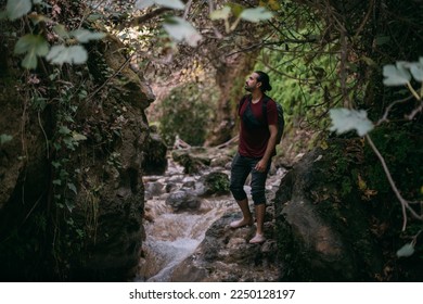 A young male tourist walks along a shallow tropical mountain river among the rocks.  A man with a backpack walks between rocks overgrown with tropical plants.   - Shutterstock ID 2250128197