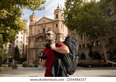 A young male traveler and backpacker, wearing sunglasses, enjoys and observes the surroundings, during a sightseeing tour in a Spanish city.