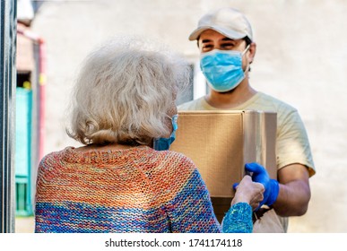 Young Male Volunteer In Mask Gives An Elderly Woman Boxes With Food Near Her House. Son Man Helps A Single Elderly Mother. Family Support, Caring. Quarantined, Isolated. Coronavirus Covid-19. Donation