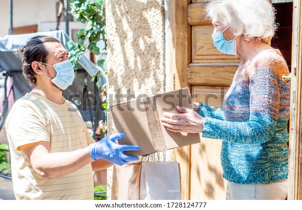Young male volunteer in mask gives an elderly\
woman boxes with food near her house. Son man helps a single\
elderly mother. Family support, caring. Quarantined, isolated.\
Coronavirus covid-19.\
Donation