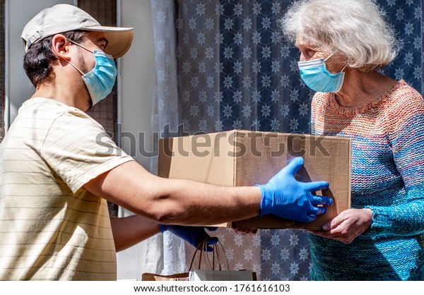 Young male volunteer in mask gives an elderly\
woman boxes with food near her house. Son helps a single elderly\
mother. Family support, caring. Quarantined, isolated. Coronavirus\
covid-19. Donation