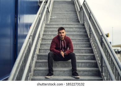 A young male wearing a red jacket with dark jeans and sitting on the steps outdoors - Shutterstock ID 1852273132