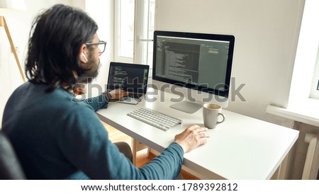 Young male web developer working from home, using desktop computer and laptop, writing code. Freelance, working online, home office concept. Focus on left hand. Stay home, self isolation