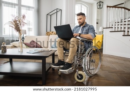 Young male wheelchair user in casual clothes looking at laptop screen during online work in modern living room. Concept of technology and remote job for people with disabilities.