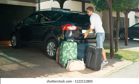 Young Male In White Shirt Closes The Trunk Of Black Car Parked In Front Of His House In The Sunny Suburbs. Caucasian Man Finishes Unpacking His Luggage In The Driveway And Shuts The Boot Lid Of SUV.