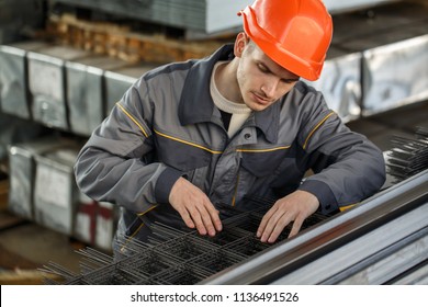 Young male worker wearing orange hardhat and gray protective suit checking quality of material on metal stock. Concentrated man leaning over metal sheet and looking at it. Metal pallets on background.