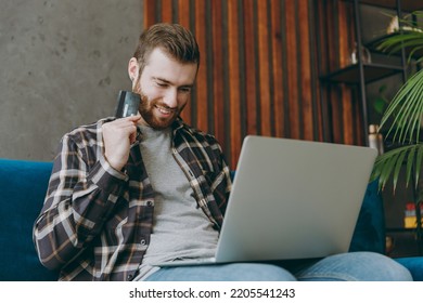 Young Man 20s He Wear Brown Shirt Using Laptop Pc Computer Hold Credit Bank Card Shopping Online Order Delivery Book Tour Sit On Blue Sofa In Living Room Apartment Stay Home Indoor Flat On Weekends.