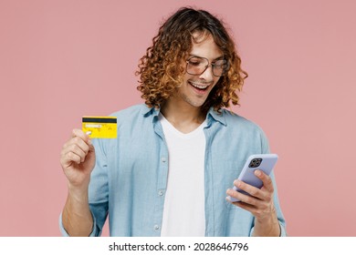 Young Man 20s With Long Curly Hair In Blue Shirt Glasses Using Mobile Cell Phone Hold Credit Bank Card Doing Online Shopping Order Delivery Booking Tour Isolated On Pastel Plain Pink Background Studio