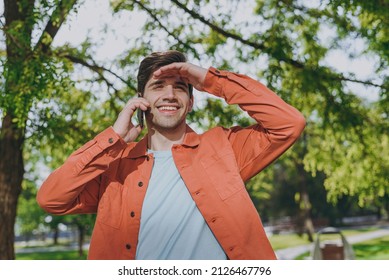 Young Man 20s In Orange Jacket Walk Talk Speak On Mobile Cell Phone Look Far Away Distance Rest Relax In Spring Green City Park Go Down Alley Sunshine Lawn Outdoors On Nature. Urban Leisure Concept