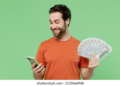 Young man 20s wear casual orange t-shirt holding fan of cash money in dollar banknotes mobile cell phone isolated on plain pastel light green color background studio portrait. People lifestyle concept