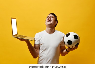 young man of the 20s in a white T-shirt, holding a laptop and a soccer ball, clung to the screen - closely watching watching the transfer on the screen. worried - Shutterstock ID 1408029638