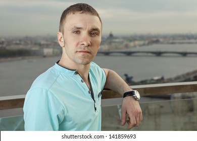 A Young Man 27 Years Old, Caucasian, Slav, Russian, Dressed In Turquoise Shirt With Short Sleeves, Horizontal Portrait, Close-up, Against Background Of Panorama Of European Cities. One Person, Male.