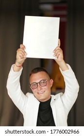 A Young Man Of 30-35 Years Old In A White Jacket, Black Turtleneck, Glasses And A Short Haircut Holds An Empty Sign In His Hands. Template For Design, Mockup.