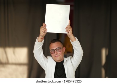 A Young Man Of 30-35 Years Old In A White Jacket, Black Turtleneck, Glasses And A Short Haircut Holds An Empty Sign In His Hands. Template For Design, Mockup.