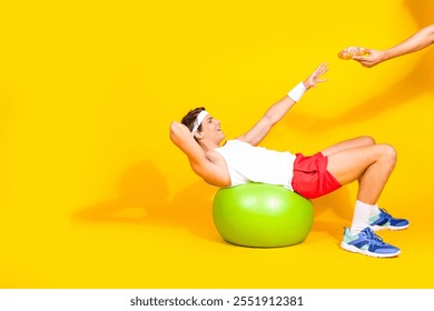 Young man in activewear balancing on fitness ball against yellow background reaching for donut - Powered by Shutterstock