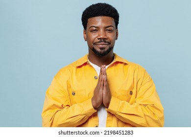 Young Man Of African American Ethnicity 20s In Yellow Shirt Hold Hands Folded In Prayer Gesture, Begging About Something Isolated On Plain Pastel Light Blue Background Studio. People Lifestyle Concept