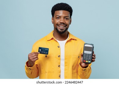 Young man of African American ethnicity 20s wear yellow shirt hold wireless modern bank payment terminal to process acquire credit card payments isolated on plain pastel light blue background studio.