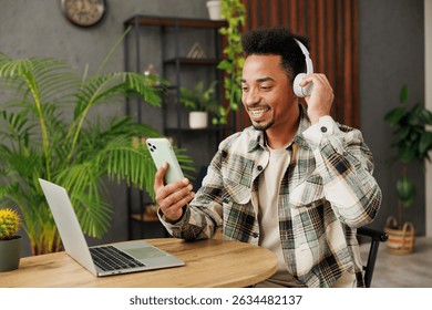 Young man of African American ethnicity wear casual clothes listen to music in headphones use mobile cell phone sitting at table in coffee shop cafe relax rest in restaurant during free time indoors