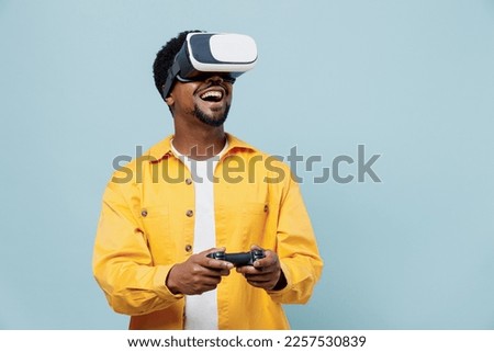 Young man of African American ethnicity 20s wear yellow shirt hold in hand play pc game with joystick console watching in vr headset pc gadget isolated on plain pastel light blue background studio.