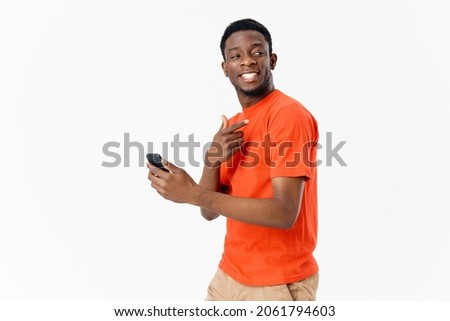 young man of african appearance with a mobile phone and in an orange t-shirt on a light background cropped view