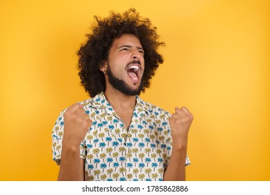 Young man with afro hair over  wearing hawaiian shirt standing over yellow background celebrating surprised and amazed for success with arms raised and eyes closed. Winner concept. - Shutterstock ID 1785862886