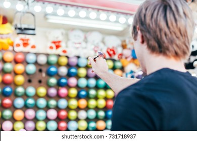 Young man aims at wall of colourful air balloons in order to win teddy bear or plush toy prize for his girlfriend, during romantic date at amusement park or carnival