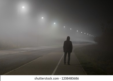 Young man alone slowly walking under white street lights in night. Dark time. Peaceful atmosphere in mist. Foggy air. Back view.