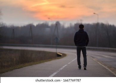 Young man alone slowly walking on sidewalk in dark evening. Orange sunset light in sky. Back view.