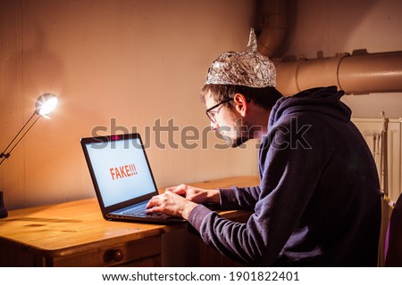 Young man with aluminum cap is sitting in the dark basement in front of a laptop. Conspiracy theory concept