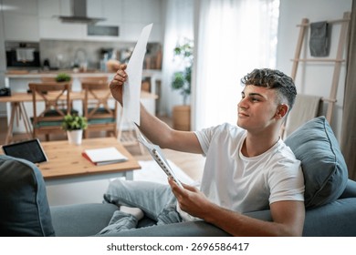 Young man analyzing documents and paperwork while sitting comfortably on a couch in a modern living room, focusing on his studies or personal finances