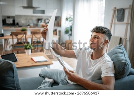 Young man analyzing documents and paperwork while sitting comfortably on a couch in a modern living room, focusing on his studies or personal finances