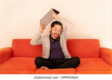 Young Man Angry With Rage Throwing The Computer, Sitting On An Orange Couch Sofa At Home In The Living Room. Stress Concept.