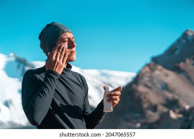 A young man applies sunscreen to his face in the highlands, in the background the peaks of the mountains are covered with snow. The traveler takes care of his skin while climbing.
