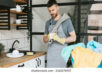 young man in apron and gloves does household chores, washes dishes with towel on his shoulder, men housework, household help in stylish kitchen in modern apartment