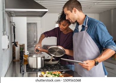 Young Man In Apron Helping African Woman To Prepare Lunch. Happy Girl Adding Salt In Pot For Pasta While Guy Hold The Lid Up. Multiethnic Couple Cooking Together With The Help Of A Digital Tablet.