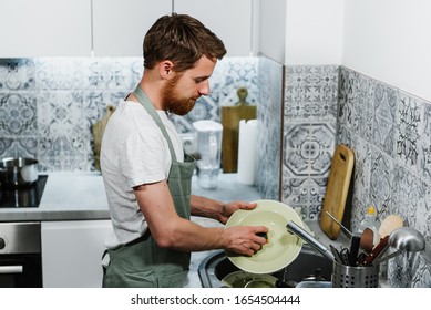 A young man in an apron washes dishes in the kitchen. - Powered by Shutterstock