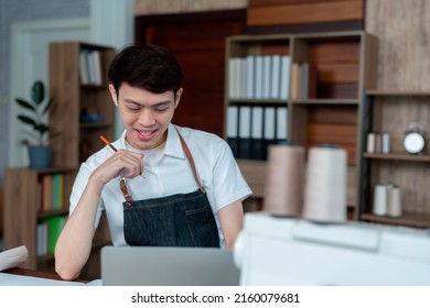 Young Man Asian LGBTQ Tailor Shop Owner, A Designer Sits Designing A Set That Must Be Cut On A Large Sheet Of Paper On A Table.Small Business Professional Design Clothes To Sell Online.