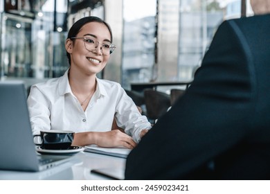 Young man and asian woman discussing work over a cup of coffee sitting at business center cafe, using laptop computer. Businessman and businesswoman partners having a meeting in office lobby. - Powered by Shutterstock