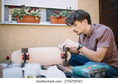 Young man assembling skateboard while sitting outdoor. Concentrated man fitting up skateboard wheels to wooden board in yard. - Powered by Shutterstock