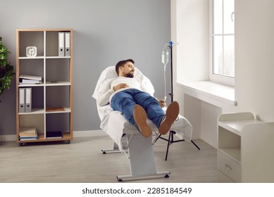 Young man attached to intravenous drip lying on bed in medical clinic. Male patient getting vitamin IV infusion drip therapy in hospital room. Medical treatment, maintenance of immune system - Shutterstock ID 2285184549