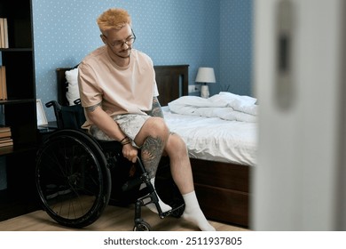 Young man attending to activities in a bedroom while seated in a wheelchair, with tattoos on arm and glasses on face, with some personal belongings and a bed in background - Powered by Shutterstock