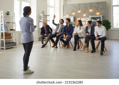 Young Man In Audience Raises Hand To Ask Speaker A Question. Small Group Of People Sitting In Row In Modern Office Space Listening To Business Trainer, Corporate Psychologist Or Personal Growth Coach