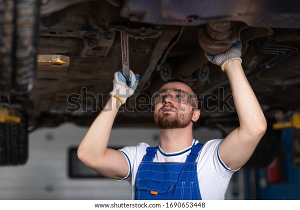A\
young man auto mechanic in overalls at his workplace repairs the\
car\'s suspension, shines a lantern on the chassis. Concept Worker\
at an automobile repair shop, a car lifted on a\
lift