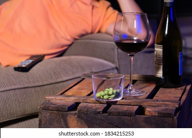 Young Man, Bachelor Sitting On A Couch, Drinking Wine And Eating Unhealthy Snacks, And Watching Movie, Series, Tv Show In The Late Evening, Night.