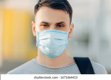 A young man in the background of an open- air building wearing a medical face mask that protects against the spread of coronavirus disease. Close- up of a man with a surgical mask on his face