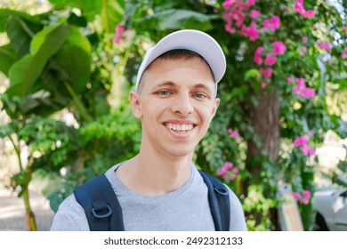 Young man with a backpack and cap smiling happily in a park. Natural outdoor setting with green foliage and pink flowers. Concept of joy and travel - Powered by Shutterstock